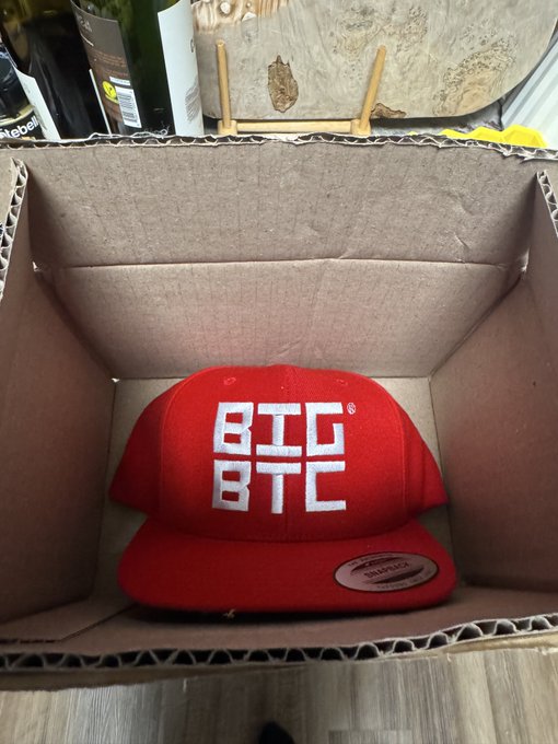 A red snapback hat with a white 'BTC BTC' logo inside an open cardboard shipping box, with liquor bottles and a wooden stand visible in the background.