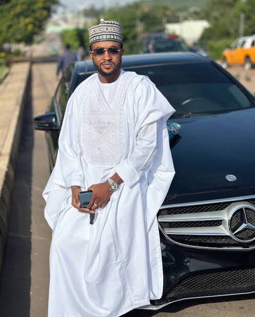 A man in traditional white attire and embroidered cap stands beside a black Mercedes-Benz, holding a phone and smiling at the camera. The image appears to introduce or honor @Gayu_BTC, who is being acknowledged in the accompanying post as someone who provided help during a difficult time.