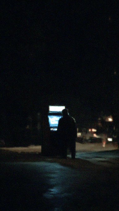 A person stands silhouetted next to a brightly lit phone booth on a dark street at night.
