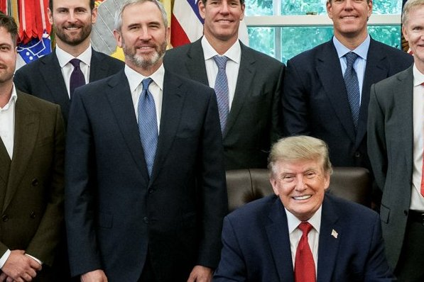 President Donald Trump sits at his desk in the Oval Office smiling broadly, surrounded by a group of men in business suits posing for a formal photograph. The image appears to be from an official White House meeting, though it has no direct connection to the post's content about XRP, Brad Garlinghouse, or SEC/CFTC crypto rulemaking.