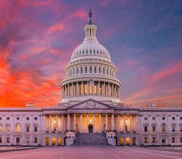 A scenic photograph of the United States Capitol building in Washington D.C., illuminated at sunset with a vibrant sky of orange, pink, and purple clouds behind its white dome.