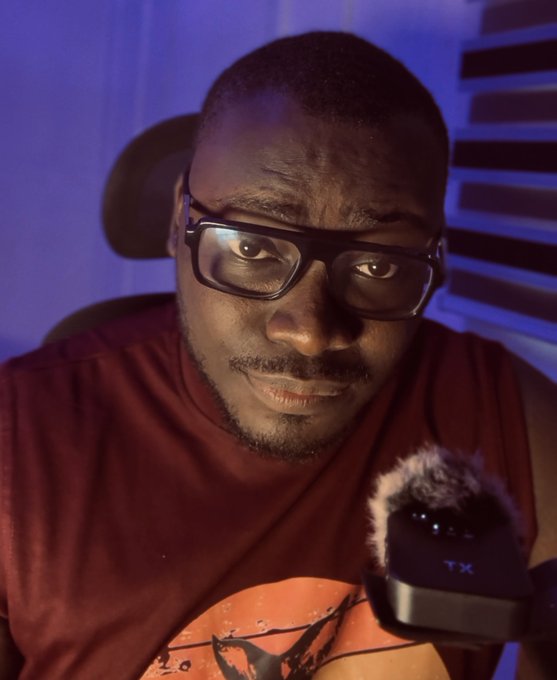 A young Black man wearing rectangular glasses and a maroon sleeveless shirt leans forward with a serious expression, with a black microphone visible in the foreground against a backdrop of blue-lit window blinds.

The image, paired with the post about market opportunities and alpha groups, suggests the subject is a content creator or streamer promoting his involvement in trading, crypto, and meme/NFT investments to his audience.
