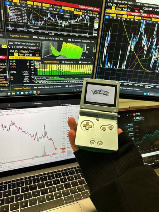 A hand holds a Game Boy Advance SP displaying the Pokémon logo, set against a backdrop of multiple monitors showing stock charts with tickers like HSY, HSY US, and YHOO, alongside a laptop displaying BTC charts.