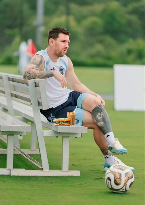 Lionel Messi in Argentina training gear sitting on a bench with a sandwich and iced coffee beside him, a soccer ball at his feet on a training pitch.