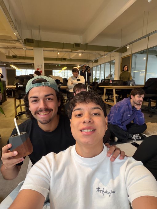 Two young men taking a selfie in an office space, with the man on the left holding a drink and wearing a backwards cap, and the man on the right wearing a white sweatshirt with an 'artcollective' logo.