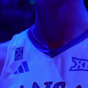 A close-up of a person wearing a white Adidas XRP basketball jersey with 'XII' visible on the chest, illuminated by dark blue and purple lighting.