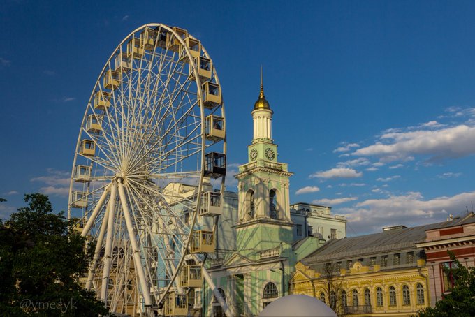 The image features a large white Ferris wheel with gold accents against a clear blue sky. The Ferris wheel is situated next to an old building, possibly a church or another historical structure. In the background, there are trees that add greenery to the scene. The photo also includes text about GM CT and Season 5 of "River Points" on the right side.