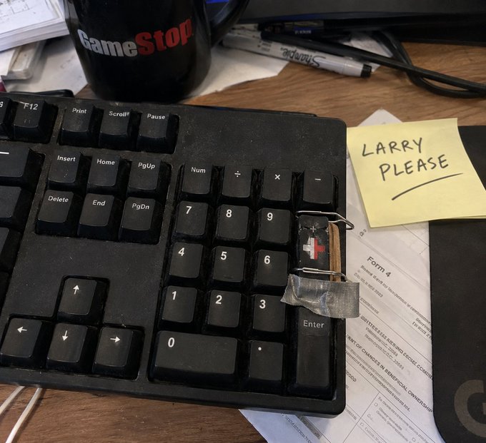 A worn keyboard sits on a cluttered desk next to a sticky note reading "LARRY PLEASE," a GameStop mug, and tax forms, with the plus key duct-taped onto a paperclip repair—capturing the "Larry, we need to talk about your keyboard" post perfectly.