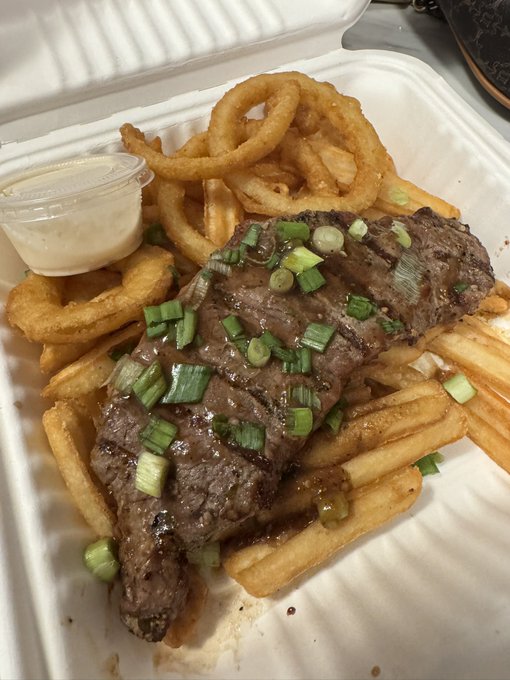 A sizzling steak topped with chopped green onions rests on a bed of golden french fries, served with onion rings and a side of dipping sauce in a styrofoam takeout container. The hearty meal perfectly satisfies the bold appetite of a hungry Leo, ready to devour this protein-packed plate.