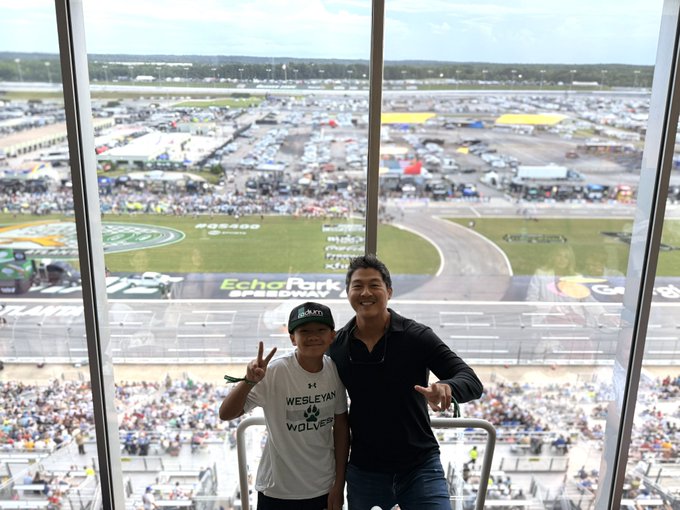 A father and son pose with peace signs in a stadium suite overlooking a NASCAR race track, with "EchoPark Speedway" and "Goodyear 400" visible on the track and infield.