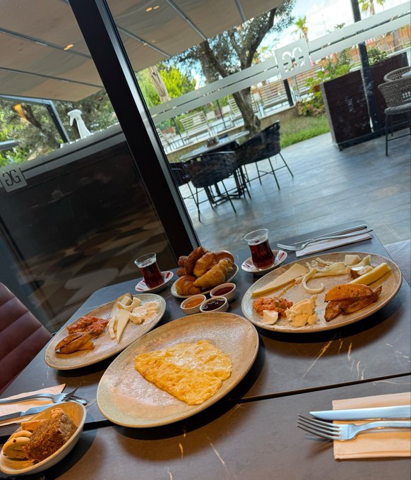 A table at a restaurant with an outdoor view, featuring three plates of food including a bread basket, cheese, omelet or pancake, tea in a traditional glass, and various spreads and jams, with cutlery and napkins arranged.