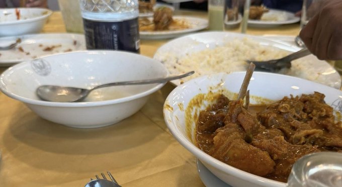 The image shows a dining table with various dishes, including bowls filled with food such as rice and curry. A person can be seen eating from one of these bowls using a fork and knife. The table also has other items like cups, spoons, forks, and knives scattered around it. In the background, there is another person partially visible who appears to be engaged in conversation or preparing their meal. The overall atmosphere suggests a casual dining experience with friends or family gathered together for a meal.