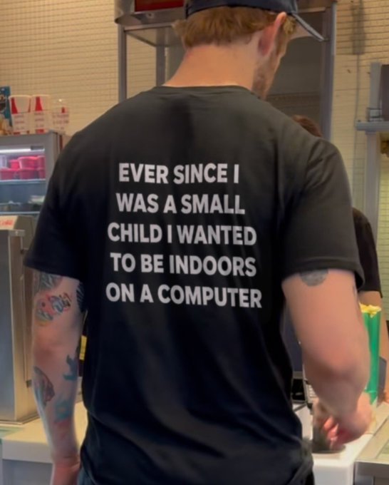 A man with tattoos and a backward cap is photographed from behind wearing a black t-shirt that reads: 'EVER SINCE I WAS A SMALL CHILD I WANTED TO BE INDOORS ON A COMPUTER'.