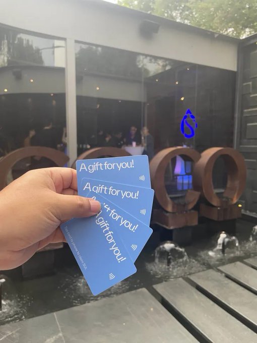 A hand holds three light blue gift cards reading 'A gift for you!' with a contactless payment symbol, held in front of a glass building with circular water fountains.