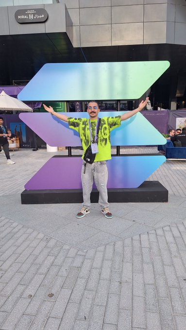 A man in a green tie-dye shirt and sunglasses poses with arms outstretched in front of a large iridescent geometric sculpture outside the 'MIRAL' building.
