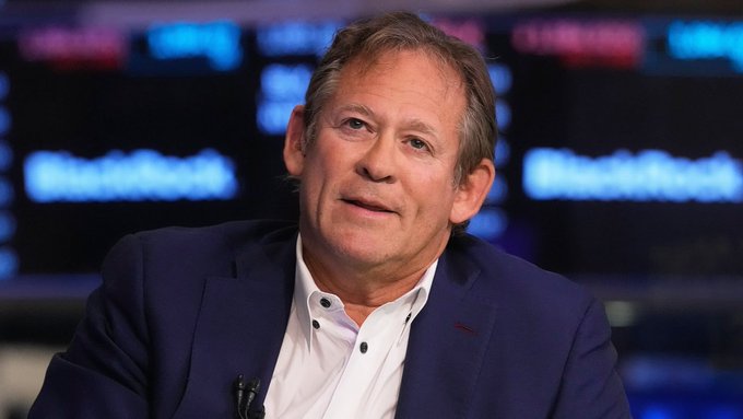 A man in a navy blazer and white shirt speaks on what appears to be a Bloomberg TV panel, with a small lapel microphone visible. The blurred studio backdrop features repeating "Bloomberg" signage, tying the image to Rick Rieder's televised commentary about Bitcoin replacing gold as a store of value.