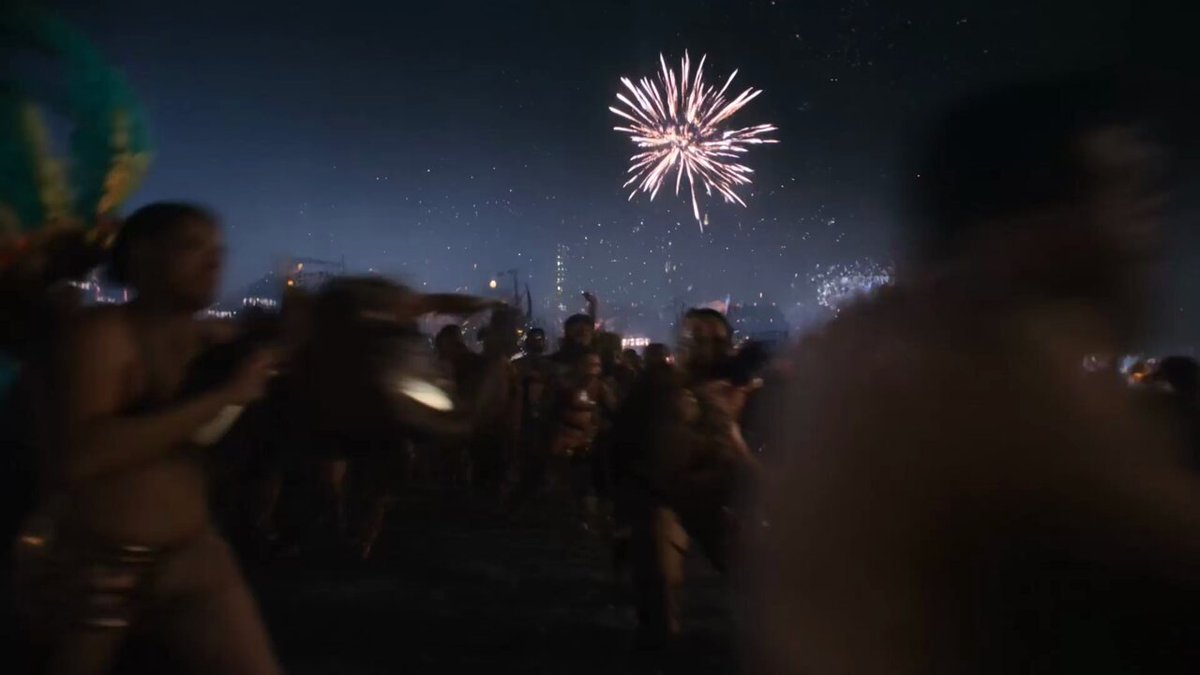 The image shows a group of people standing outside at night, watching fireworks explode above them. There are several people scattered throughout the scene, some closer together while others stand alone or with friends. A few individuals can be seen holding handbags as they enjoy the spectacle. The fireworks display is the main attraction in this outdoor gathering, creating an atmosphere of excitement and celebration for everyone present.