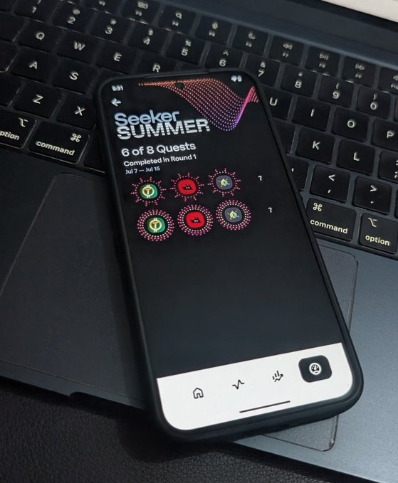 A smartphone resting on a laptop keyboard displays the "Seeker SUMMER" quest page showing "6 of 8 Quests Completed in Round 1" with several round icons, some completed.