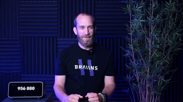 A bearded man in a black t-shirt with 'BRAIN' printed on it stands in front of blue acoustic panels, with a digital counter displaying '956 080' beside him.