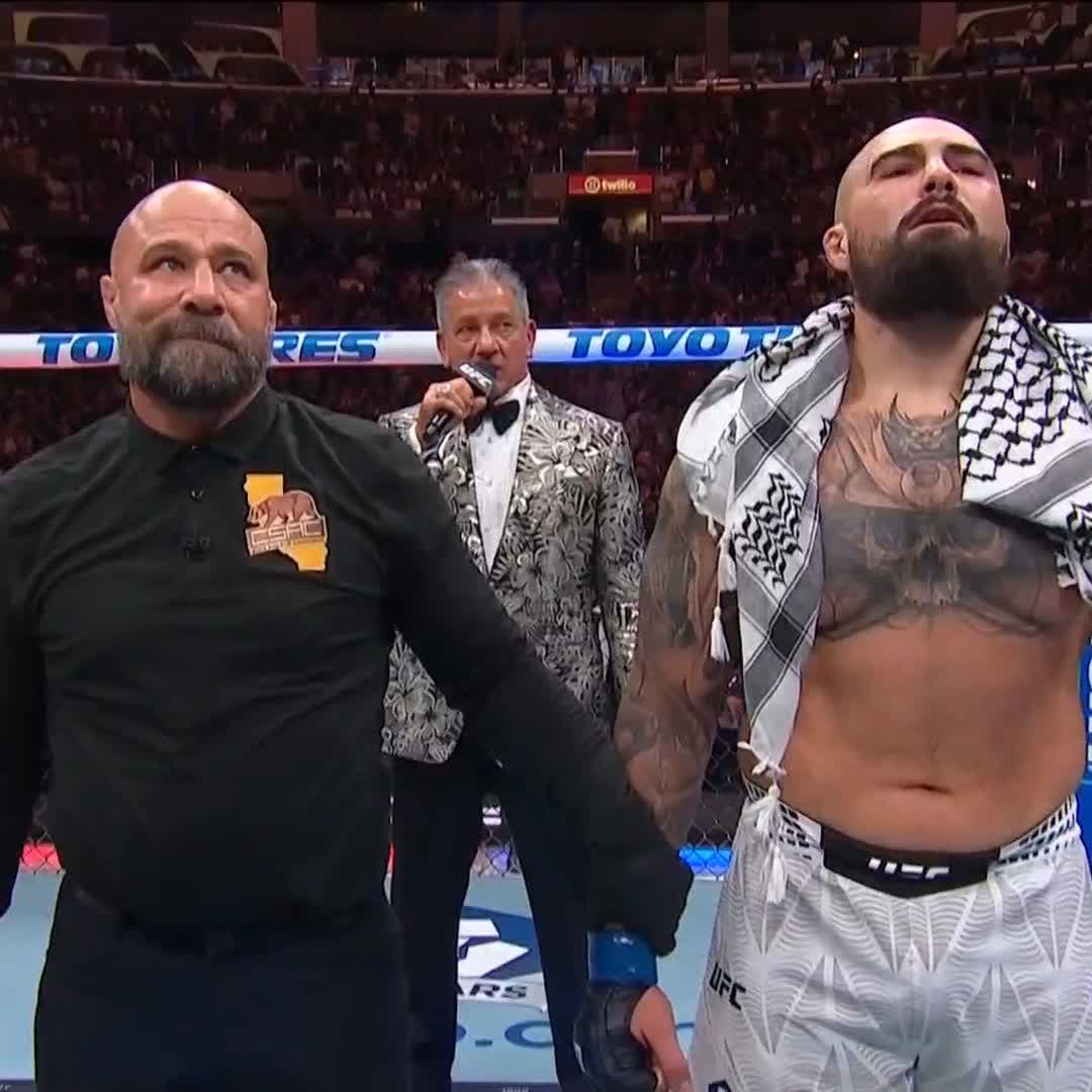 Inside a packed UFC arena with visible "TOYO TIRES" and "Twitch" signage, a shirtless, bearded fighter wearing a black-and-white keffiyeh and UFC shorts stands beside a bearded coach in a black shirt featuring a bear and California logo, while a man in a patterned tuxedo jacket holds a microphone in the background.