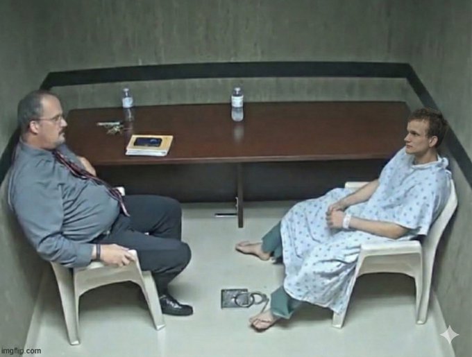 A scene from a movie shows two men sitting opposite each other in a stark, concrete-walled interrogation room with a small table between them; one wears a suit and glasses, while the other wears a hospital gown.