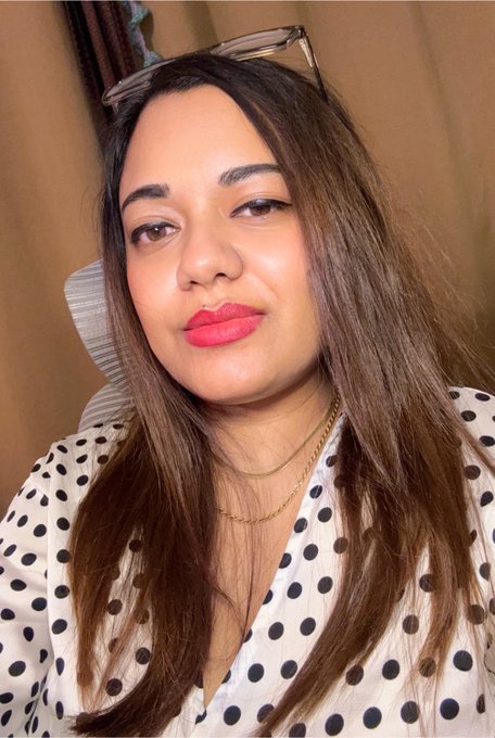 A selfie of a woman with long brown hair, pink lipstick, and a polka dot blouse, with glasses perched on her head.