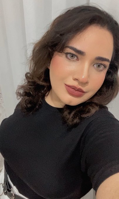 A woman with long dark wavy hair poses for a selfie, wearing a black top and sporting red lipstick, with a white curtain visible in the background.