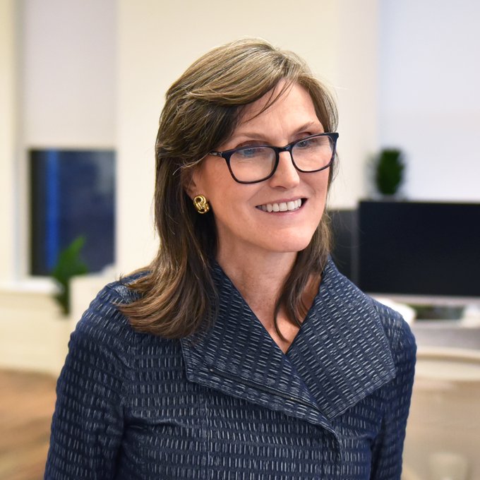 A smiling woman with shoulder-length hair, glasses, and a blue patterned blazer is pictured in what appears to be an office setting. The image accompanies news about Cathie Wood's Ark Invest purchasing $15.4M worth of Block shares amid a stock dip.
