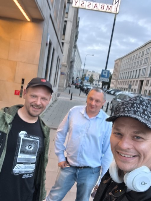 Three men take a selfie on a city street, with the man in the center wearing a light blue button-up shirt, while 'EMBASSY' signage is visible upside-down in the background.