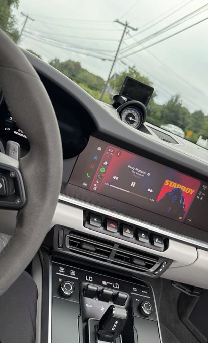 Interior view of a Porsche vehicle, showing the steering wheel with the 'PORSCHE' logo, the dashboard screen playing a song titled 'Party Moment' by Star Boy, and the center console with climate controls and gear shifter.