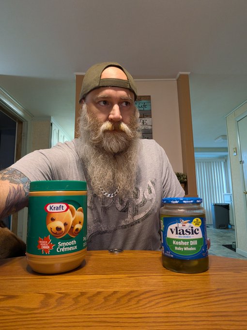 A bearded man wearing a backwards cap sits at a table displaying a jar of Kraft Smooth Cremteur peanut butter and a jar of Vlasic Kosher Dill Baby Wholes pickles.
