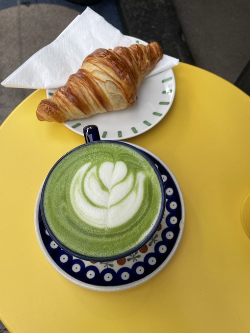 A matcha latte with leaf latte art sits in a blue and white patterned cup on a bright yellow table, accompanied by a golden croissant on a white plate with green accents.