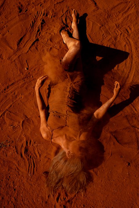 An artistic, high-contrast photograph of a woman lying on red sand or dirt, with reddish dust scattered across her body and blonde hair.