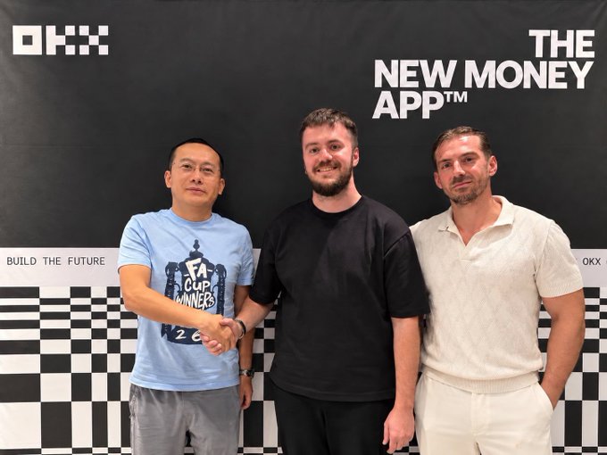 Three men pose together in front of a black backdrop featuring the "OKX" logo and "THE NEW MONEY APP™" tagline, with one wearing an FA Cup Winners shirt, appearing to shake hands as part of an OKX team and partner gathering.