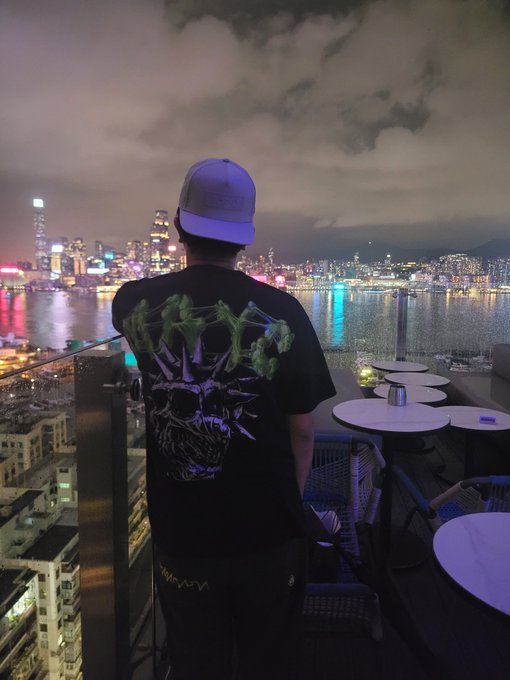 A person seen from behind, wearing a white cap and a black t-shirt with a green skull design, looking out over the Hong Kong city skyline at night from a rooftop terrace.