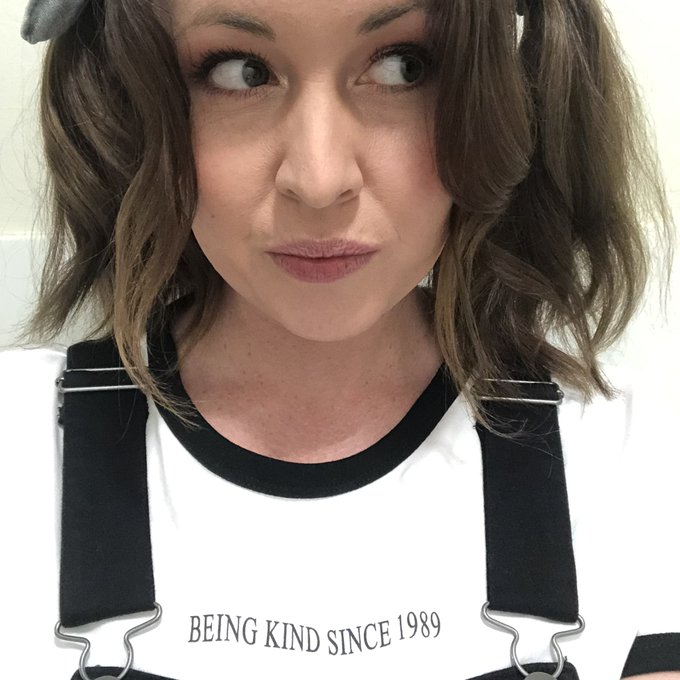 A close-up of a woman with brown hair looking to the side, wearing a white t-shirt with black overalls straps that reads 'BEING KIND SINCE 1989'.