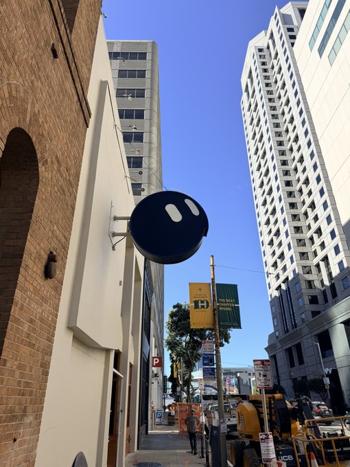 An urban street scene featuring a black sign with two white pill-shaped marks resembling a stylized face, mounted on a building with high-rises in the background.