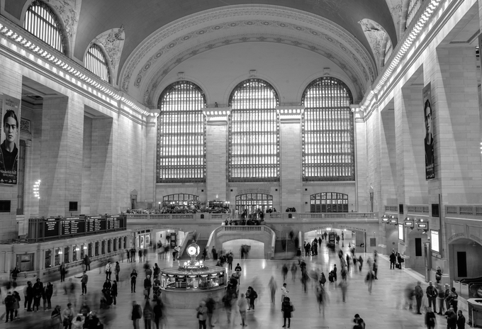 Black and white photograph of Grand Central Terminal's main concourse, showing the arched windows, the iconic clock atop the information booth, and crowds of people.