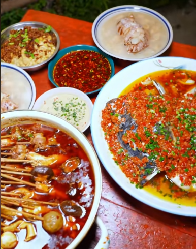 A spread of spicy Chinese dishes featuring a whole fish head topped with red chilies, skewers in red broth, and various dipping sauces arranged on a red table.