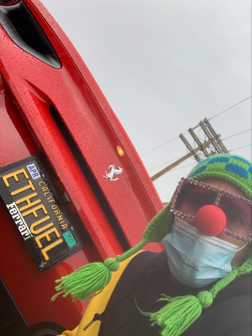A selfie of a person wearing a green knit cap, red nose, and face mask, standing next to a red Ferrari with an 'APR CALIFORNIA Ferrari' license plate.