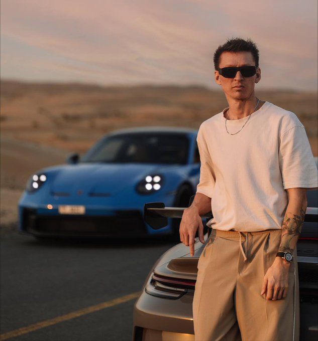 A photograph of a man in a white t-shirt and sunglasses leaning on a gold car, with a blue sports car parked behind him against a desert sunset.