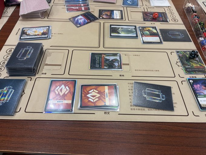 Various trading cards are laid out on a playing mat with designated slots for a card game, along with stacks of face-down cards and a few dice.