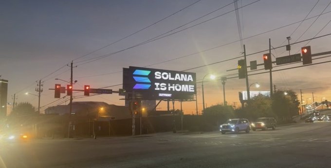 A sunset photograph of a billboard reading 'SOLANA IS HOME' with the Solana logo at an intersection with traffic lights and cars.