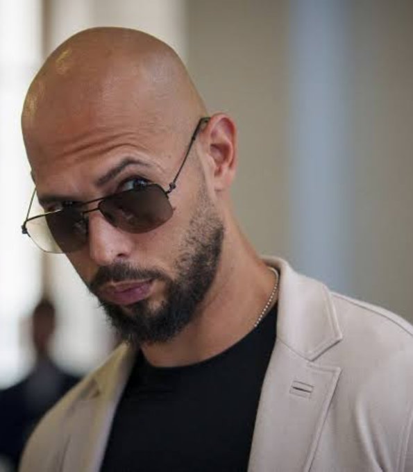 A bald, bearded man wearing aviator sunglasses and a cream-colored blazer over a black shirt looks to the side, matching the stylish persona of Andrew Tate, whose recent $1.87M sale of HYPE tokens to Binance is highlighted in the caption.