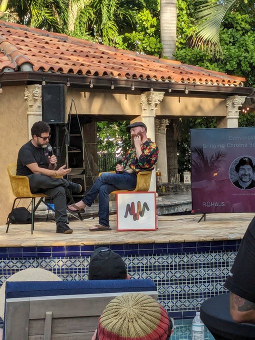 Two men sit in yellow chairs on a stage having a conversation, with one holding a microphone and the other listening; a banner behind them reads 'Conversation Bringing Chrome to' and 'ROHAUS'.