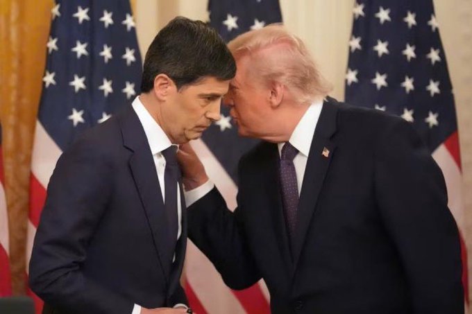 Two men in dark suits (Paul Ryan and Donald Trump) stand closely together with American flags in the background, appearing to have a private conversation.