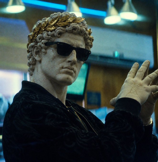 A cinematic shot of a classical marble statue adorned with a golden laurel wreath and wearing black sunglasses and a black jacket, posing with raised hands in a modern setting.
