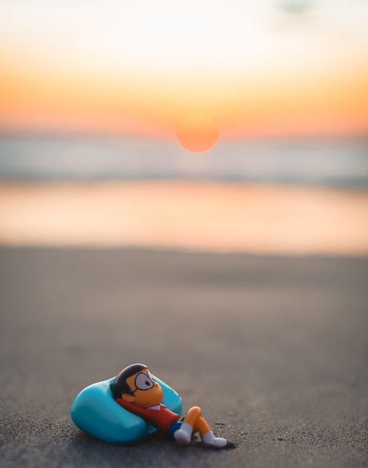 A small figurine of a cartoon character relaxing on a blue pool float on a sandy beach with a sunset over the ocean in the background.