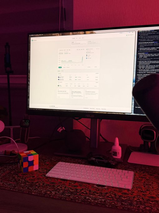 A computer desk setup with pink ambient lighting, displaying an ASUS monitor showing a dashboard, accompanied by a pink keyboard, a Rubik's cube, and other accessories on a patterned mat.