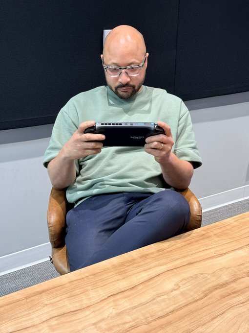 A photo of a bald man with glasses wearing a sage green t-shirt and dark pants, sitting in a tan office chair while intently using a handheld gaming device behind a wooden desk.
